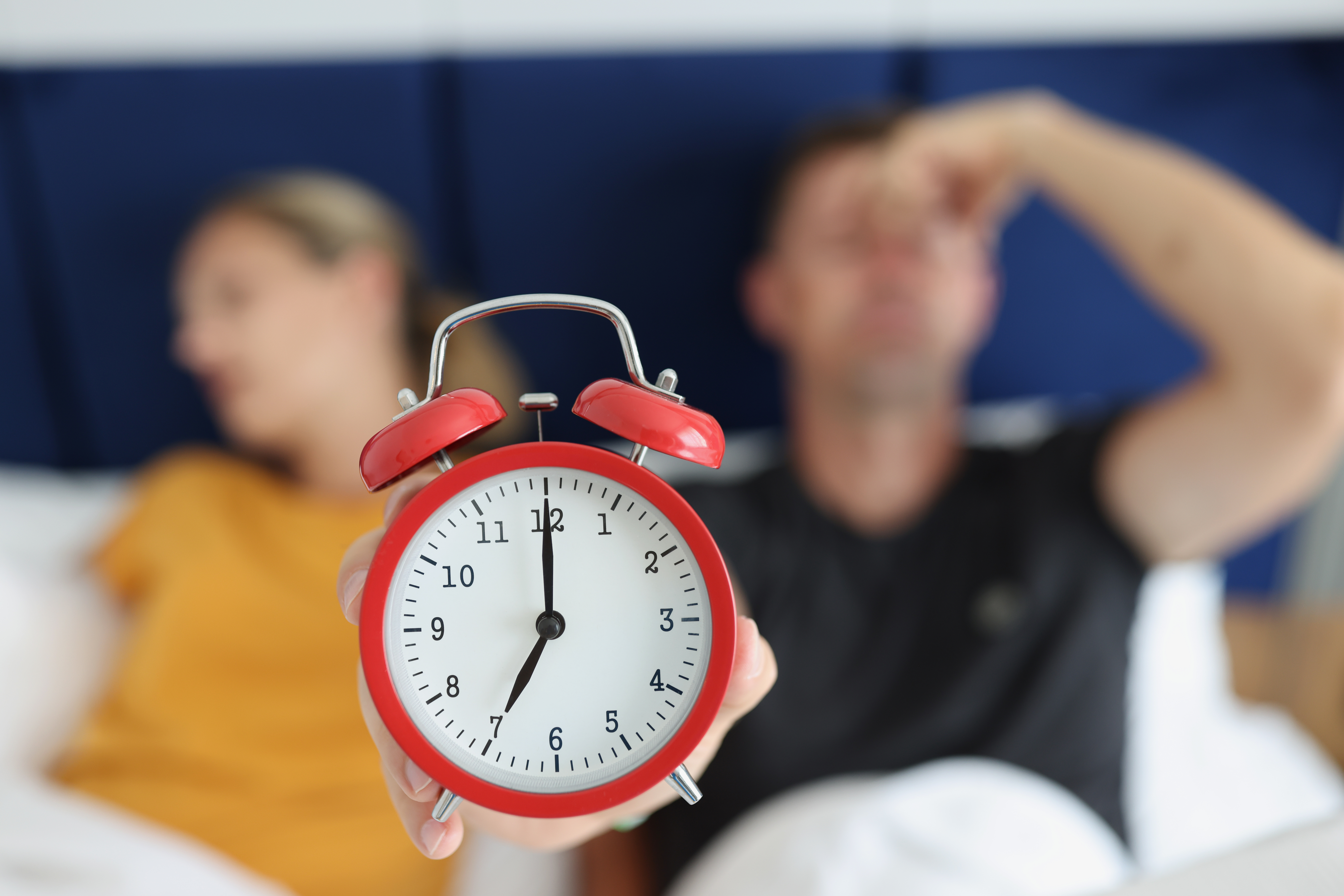 Red alarm clock against the background of a sleeping couple in a hotel, close-up, blurry. Early call, noise in the room